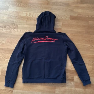 Svart hoodie från Plein Sport - Svart hoodie från Plein Sport med dragkedja framtill, huva och fickor. Snyggt röd Plein Sport-tryck på ryggen och diskret logga på dragkedjan. Tillverkad i mjuk bomull, perfekt för en sportig och avslappnad look.