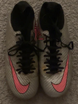Nike fotbollsskor silver/rosa - Säljer ett par Nike fotbollsskor i silver med stora rosa swoosh-loggor och svarta snören. Skorna har en låg profil och dobbar i rosa och svart för bra grepp på planen. Tillverkade i syntetmaterial för lätt känsla och bra bollkontroll. Det finns en gul fläck på framsidan men det syns inte så mycket, annars är det perfekt 
