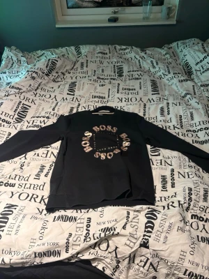 Svart sweatshirt från Hugo Boss - Säljer en svart sweatshirt från Hugo Boss med stort BOSS-tryck i beige och vitt på bröstet. Tröjan har rund halsringning, långa ärmar och ribbade muddar. Perfekt för dig som vill ha en clean och stilren look med en touch av logga. Funkar till S/M
