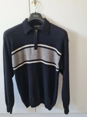 Stickad ulltröja med dragkedja från KappAhl strl S - Säljer en mörkblå stickad tröja från KappAhl med fjärdedels dragkedja och krage. Tröjan har en grå och vit rand över bröstet och långa ärmar. Materialet är en blandning av lammull 80% och polyamid 20%. Storlek S. Kan hämtas i Vallentuna.