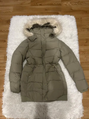 Pufferjacka med fuskpäls från Yessica - Säljer en grön pufferjacka från Yessica med avtagbar huva och fuskpäls. Jackan har dragkedja, tryckknappar och två snedställda fickor med dragkedja. Perfekt för kalla dagar och har en skön, vadderad känsla.