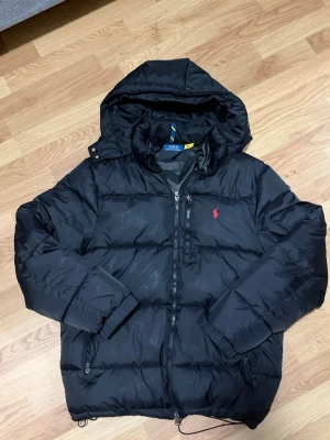 Svart pufferjacka från Polo Ralph Lauren - Säljer en svart pufferjacka från Polo Ralph Lauren med huva och dragkedja. Jackan har en klassisk röd logga på bröstet, flera fickor och justerbar nederkant med snörning. Perfekt för kalla dagar och riktigt skön att bära.