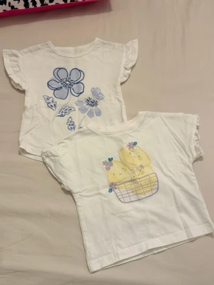 2-pack vita t-shirts Carter's 12m - Två söta vita t-shirts från Carter's i storlek 12 månader. Ena t-shirten har blå blommor broderade framtill och den andra har ett gulligt tryck med gula kycklingar i en korg och små blommor. Båda har korta ärmar med volangkant och är gjorda i mjuk bomull.