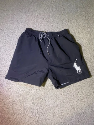 Svart badshorts Polo Ralph Lauren XL - Snygga svarta badshorts från Polo Ralph Lauren i storlek XL. Shortsen har en vit broderad logga på benet, elastisk midja med snörning och innerfoder i mesh. Perfekta för stranden eller poolen. Storlek är XL. Kan diskuteras för ett lägre pris.