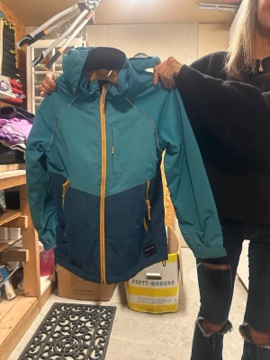 Blå vindjacka från Polarn O. Pyret - Snygg tvåfärgad vindjacka i turkos och mörkblått från Polarn O. Pyret. Jackan har huva, gula dragkedjor och praktiska fickor framtill. Perfekt för blåsiga dagar och outdoor-häng. Reflekterande detaljer och justerbara ärmslut för extra funktion.