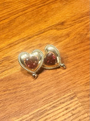 Guldörhängen med röd hjärtsten - Hjärtformade örhängen i guldton med röd sten. Oanvända, i nyskick. Originalask medföljer. Ordinarie pris 499 kr, säljes för 200 kr