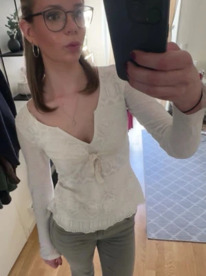 Vit Odd Molly blus med spets och knyt - Supersöt vit blus från Odd Molly med v-ringning, spetsdetaljer och ett litet knyt framtill. Långärmad och tillverkad i mjuk bomull med broderade mönster. Perfekt för dig som gillar romantisk och feminin stil.