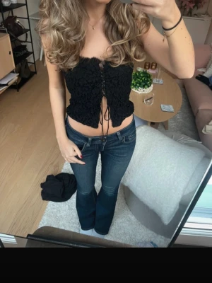 Svart croppad korsettopp med snörning - Säljer en svart croppad korsettopp med smala axelband och snygg snörning framtill. Toppen har en strukturerad, blommig spetslook och sitter tight över kroppen. Perfekt för dig som vill ha en trendig och edgy vibe.