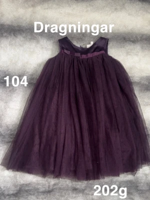 Mörklila tyllklänning med satinband - Supersöt mörklila klänning med fluffigt tyllkjol och ett matchande satinband i midjan. Ärmlös modell med rundad halsringning och flera lager tyg i kjolen för extra volym. Perfekt för dig som vill sticka ut lite extra!🌸 Dragningar i tyllen. Storlek 104