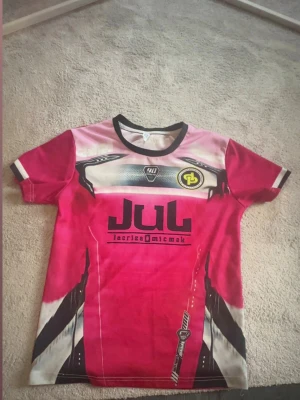 Röd sportig t-shirt JUL Ninetta - Cool röd t-shirt från JUL med svart och vit grafik, tryck framtill och texten 'NINETTA 7' på ryggen. T-shirten har svarta detaljer vid kragen och ärmarna samt sportig design med tryckta loggor och nummer. Perfekt för dig som gillar unika sportiga plagg.