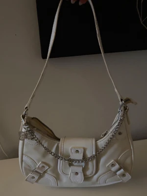 Vit axelväska med kedjedetalj - Säljer en snygg vit axelväska i syntetmaterial med silverfärgad kedja och fjärilsberlock. Väskan har spänndetaljer framtill, dragkedja upptill och en praktisk axelrem. Perfekt storlek för det viktigaste och ger en cool touch till din outfit.