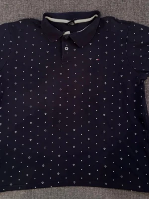 Marinblå piké från Tommy Hilfiger - Snygg marinblå pikétröja från Tommy Hilfiger med vita prickar och små ankare som mönster. Klassisk krage, kort ärm och knappslå framtill. Tillverkad i mjuk bomull som känns skön mot huden. Liten broderad logga på bröstet för extra detalj.