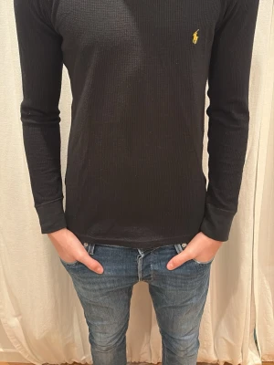 Svart långärmad tröja från Ralph Lauren - Svart långärmad tröja från Ralph Lauren med diskret ribbad struktur och den klassiska gula loggan broderad på bröstet. Tröjan har en normal passform och är tillverkad i mjuk bomull, perfekt för en clean och stilren look. Storlek S