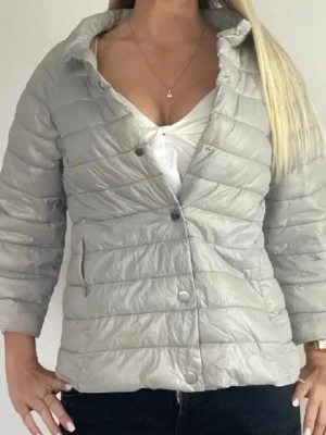 Hermo liknande jacka  - Säljer en ljusgrå pufferjacka med horisontella sömmar och tryckknappar framtill. Jackan har en hög krage och trekvartsärmar, perfekt för dig som vill ha en lätt och stilren jacka till vardags. Materialet är syntet och ger en glansig finish.