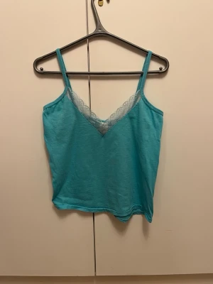 Turkos linnetopp med spets från H&M strl L / 42-44 - Turkos linnetopp från H&M i storlek L / 42-44, tillverkad i 100% bomull. Toppen har smala axelband och en snygg v-ringning med ljus spetsdetalj framtill.