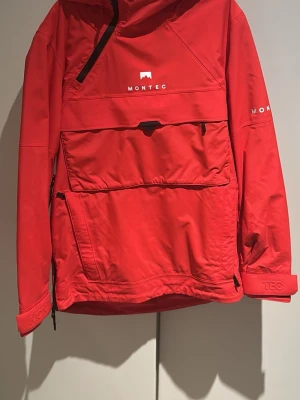 Röd vindjacka från Montec - Säljer en röd vindjacka från Montec med stor magficka, huva och dragkedjor på sidan och framtill. Perfekt skidjacka! Endast använd 1 vecka.