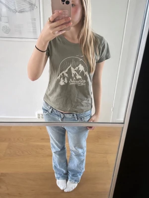 Grön t-shirt med tryck från KappAhl - Säljer en grön t-shirt från KappAhl med ett snyggt vitt tryck av berg, träd och texten 'Adventure is calling' på bröstet. T-shirten har rund halsringning och korta ärmar. Perfekt för dig som gillar outdoor vibes och enkel stil.