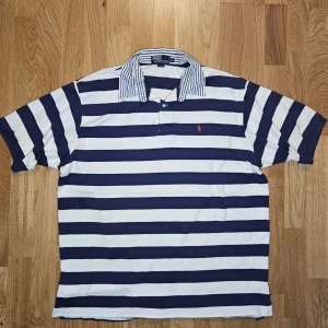 Randig pike från  Polo Ralph Lauren - Klassisk rugbyskjorta från Polo Ralph Lauren i marinblått och vitt med breda ränder och ikonisk röd logga broderad på bröstet. Skjortan har korta ärmar, knappar i halsen och en krage med smala blå ränder. Tillverkad i 100% bomull.