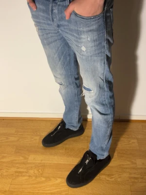Ljusblå slitna jeans med raka ben - Säljer ett par ljusblå jeans med slitningar och hål på knäna. Jeansen har raka ben, klassisk femficksdesign och coola detaljer som färgstänk och slitna partier. Perfekta för en avslappnad streetstil och passar till sneakers eller boots.