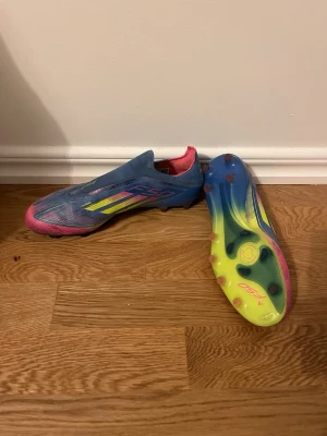 Adidas F50 blå/gul fotbollsskor - Säljer ett par Adidas F50 fotbollsskor i blått, gult och rosa. Skorna har en strömlinjeformad design utan snörning, med mönstrade detaljer och dobbar för gräsplan. Perfekta för dig som vill sticka ut på planen!