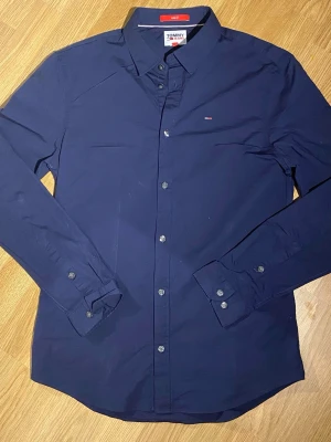 Mörkblå skjorta från Tommy Jeans - Snygg mörkblå skjorta från Tommy Jeans med klassisk krage och diskret logga på bröstet. Skjortan har långa ärmar, knappar framtill och är gjord i mjuk bomull. Perfekt till jeans eller chinos för en clean look.