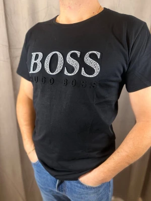 Svart BOSS t-shirt med stor logga - Snygg svart t-shirt från Hugo Boss med stor, upphöjd BOSS-logga i vitt och svart på bröstet. Klassisk rund hals och korta ärmar. Tillverkad i mjuk bomull som känns skön mot huden. Perfekt för dig som gillar stilrena och trendiga plagg.