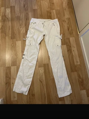 Vita cargopants med detaljer - Säljer ett par vita cargopants från AC med flera fickor, silverfärgade nitar och knappar. Storlek S 