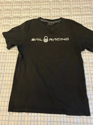 Sail Racing t-shirt Svart - Svart t-shirt från Sail Racing med vit logga och text framtill. Klassisk rund halsringning och korta ärmar. Tillverkad i mjuk bomull som känns skön mit huden. Storlek 170