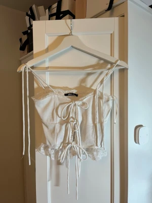 Vit snörad linnetopp Gina Tricot - Supersöt vit linnetopp från Gina Tricot med tunna axelband, ribbad struktur och snörning framtill. Toppen har spetsdetaljer längs kanterna och volang längst ner för en extra romantisk vibe. Perfekt för sommarens alla tillfällen!