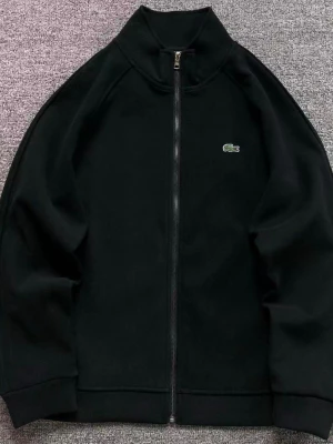 Svart zip-kofta från Lacoste - Svart kofta från Lacoste med hel dragkedja framtill och klassisk ståkrage. Liten broderad krokodil-logga på bröstet. Tröjan har långärmad design och är tillverkad i mjukt material som passar perfekt för kyliga dagar.