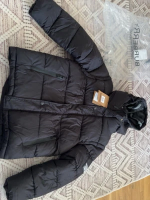 Svart pufferjacka från Burberry - Säljer en svart pufferjacka från Burberry med avtagbara ärmar så den kan användas som väst. Jackan har huva med klassiskt rutigt foder, två dragkedjefickor och tryckknappar framtill. Perfekt för kalla dagar och riktigt snygg streetstil.