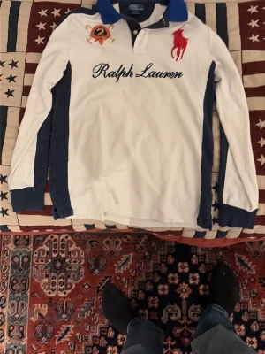  Ralph Lauren Tröja - Vit långärmad piké från Ralph Lauren med blå detaljer, stor röd logga på bröstet och broderad text. Klassisk krage och knappar framtill. 