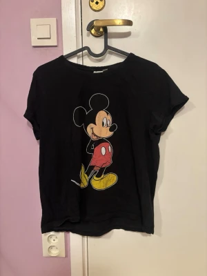 Svart Disney t-shirt med Mickey Mouse - Svart t-shirt från Disney med stort tryck av Mickey Mouse på framsidan. Klassisk rund hals och korta ärmar. Perfekt för dig som gillar ikoniska motiv och vill ha en bekväm och stilren t-shirt.