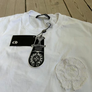 Chrome Hearts x Commes des Garçon - Säljer en vit t-shirt från Chrome Hearts med broderad logga på bröstet och detaljer på ryggen. T-shirten har rund hals och är i mjuk bomull. Perfekt för dig som gillar stilrena plagg med exklusiva detaljer.