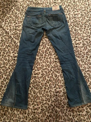 True Religion bootcut lågmidjade jeans - Säljer ett par blå bootcut, riktigt lågmidjade jeans från True Religion med ikoniska bakfickor och kontrastsömmar. Materialet är jeans i bomull med lätt slitning på benen och utställda ben för en snygg siluett. Har själv sytt om benen från skinny till bootcut.