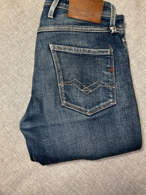 Replay Anbass jeans blå W29 L32 - Snygga Replay Anbass jeans i mörkblå/blå tvätt med snygga slitningar och fade-detaljer. Modellen har raka ben/slim fit med en modern passform, klassiska fem fickor och kontrastsömmar som ger en clean look. Läderpatch bak i midjan och Replay-detaljer på både fram- och baksida. Tillverkade i hållbar bomull för en skön känsla och stilren outfit.  Storlek: W29 L32 Modell: Replay Anbass Skick: Bra skick  Skickas samma dag vid snabb affär 📦⚡️
