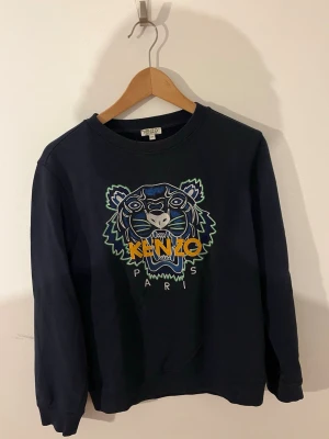 Mörkblå Kenzo sweatshirt med tiger - Säljer en mörkblå sweatshirt från Kenzo med ikoniskt broderat tigerhuvud och färgglad logga på bröstet. Tröjan har rund halsringning och långa ärmar. Perfekt för dig som vill ha en statement piece i garderoben. Använd en del gånger men fortfarande bra skick pga den fina kvaliteten. MVH SIEO Fashion 