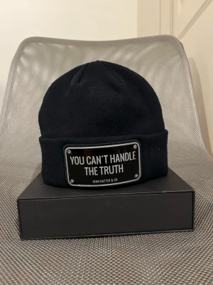Mörkblå John Hatter & Co - Säljer en mörkblå mössa från John Hatter & Co med en metallskylt framtill där det står 'YOU CAN'T HANDLE THE TRUTH'. Mössan är ribbstickad och har uppvikt kant för en clean och modern look. Använd fåtal gånger. Skickas i originaförpackningen. MVH SIEO Fashion