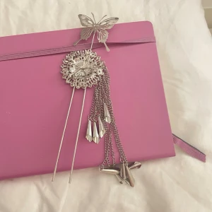Silvrig hårpin med fjäril och kedjor - Säljer en silvrig hårpin med en detaljerad fjäril på toppen och en rund, utskuren blomliknande dekoration. Flera tunna kedjor med konformade hängen hänger ner för extra effekt. Perfekt accessoar för att lyfta din frisyr. 