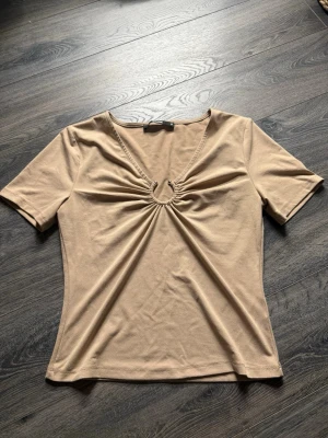 Beige topp med v-ringning och ringdetalj - Säljer en beige topp från Gina Tricot med korta ärmar och snygg v-ringning. Toppen har en ringdetalj framtill med rynkade veck som ger en cool twist. Tillverkad i mjuk bomullsmix och sitter normal på kroppen. Perfekt till jeans eller kjol!