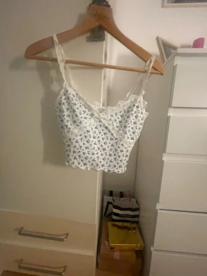 Blommig vit och blå linnetopp i st S - Supersöt linnetopp med tunna axelband och spetsdetaljer längs kanterna. Toppen är vit med små blå blommor över hela och har en croppad passform. 