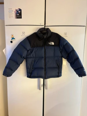 Blå och svart pufferjacka The North Face - Säljer en riktigt fet pufferjacka från The North Face i blått och svart. Jackan har hög krage, dragkedja framtill och två sidofickor med dragkedja. Perfekt för kalla dagar och har den klassiska loggan på bröstet och ryggen.