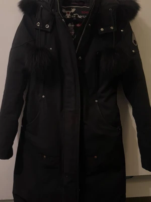 Moose Knuckles parka - Lång svart parkas äkta från Moose Knuckles med fluffig päls runt huvan samt snörningen och snygga metalldetaljer på ärmen. Jackan har yx-dragkedja. Klassisk logga på ärmen och flera fickor framtill och innerfickor. Använts väldigt sparsamt, nästintill nyskick. Priset kan diskuteras vid snabb affär.