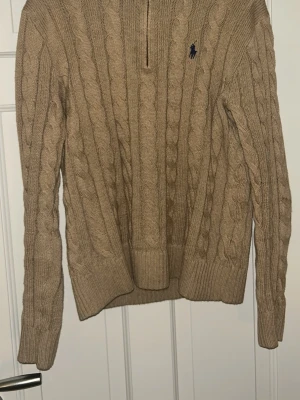 Beige kabelstickad tröja Polo Ralph Lauren - Kabelstickad beige tröja från Polo Ralph Lauren med mörkblå broderad logga på bröstet. Tröjan har lång ärm, ribbade muddar och en liten half zip vid halsen. Perfekt för lager på lager under höst och vinter.