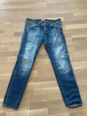 Blå slitna jeans från Jack & Jones - Säljer ett par blå jeans från Jack & Jones med snygga slitningar och tvättade detaljer. Modellen har klassisk femficksdesign, normal passform och raka ben. Jeansen är tillverkade i bomull och har en cool, avslappnad vibe.