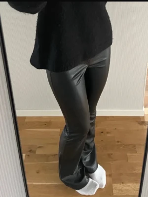 Svarta bootcut byxor i skinnimitation - Säljer ett par svarta byxor i skinnimitation med en medelhög midja och bootcut-modell. Byxorna har en snygg glansig finish och sitter tight upptill med utsvängda ben. Perfekta för dig som är runt 170cm lång. Säljs inte längre!