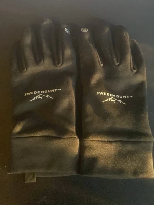 Svarta Swedemount handskar - Säljer ett par svarta handskar från Swedemount med diskret logga och bergmotiv på ovansidan. Handskarna har greppvänliga detaljer på insidan av fingrarna och är tillverkade i ett mjukt syntetmaterial. Perfekta för kyliga dagar och outdoor-aktiviteter.