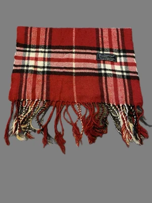 Rutig röd scarf från Burberrys - Säljer en klassisk rutig scarf från Burberrys i rött, svart och vitt. Scarfen är tillverkad i 100% lammull och har fransar längs nederkanten. Perfekt för dig som gillar tidlös brittisk stil och vill ha något extra till din outfit. Perfekt nu när snön har kommit❄️ Kom med frågor och prisförslag!