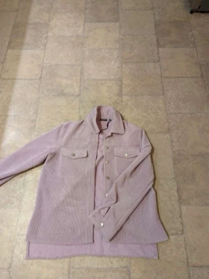 Ljusrosa manchesterjacka från Pieces - Säljer en ljusrosa overshirt i manchester från Pieces, storlek XS. Jackan har två bröstfickor med knappar och stängs med knappar framtill. Perfekt för lager-på-lager och har en avslappnad, loose passform.