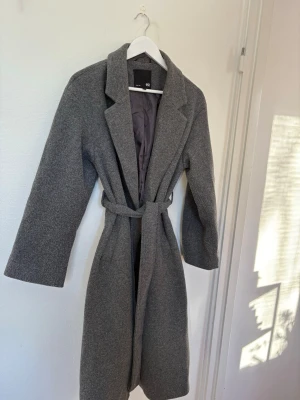 Grå kappa - Snygg grå kappa från 157 i storlek S. Klassisk modell med bred krage, knytbälte i midjan. Lite nopprig, och ett hål på insidan men inget som syns speciellt mycket. Se bilder för allmänt skick. 